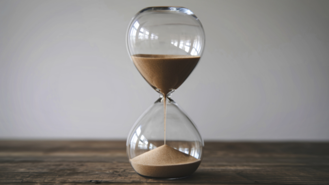 klepsydra z przesypującym się piaskiem, stoi na drewnianym stole, w tle szara ściana