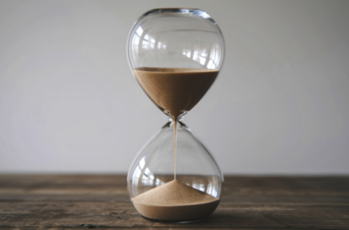 klepsydra z przesypującym się piaskiem, stoi na drewnianym stole, w tle szara ściana