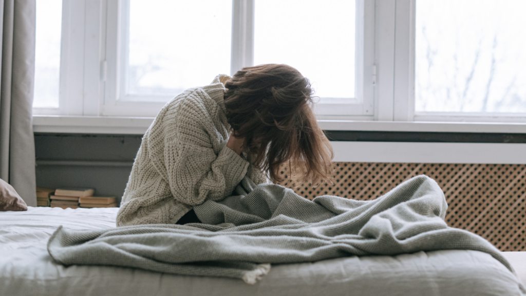 kobieta siedzi na łóżku, zakrywa twarz w dłoniach, dookoła leżą koce, w tle duże okno