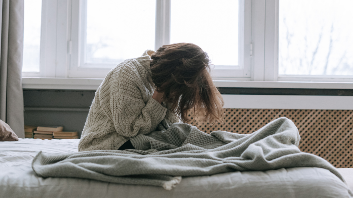 kobieta siedzi na łóżku, zakrywa twarz w dłoniach, dookoła leżą koce, w tle duże okno