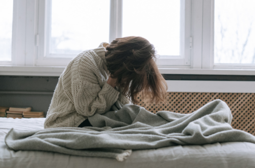kobieta siedzi na łóżku, zakrywa twarz w dłoniach, dookoła leżą koce, w tle duże okno